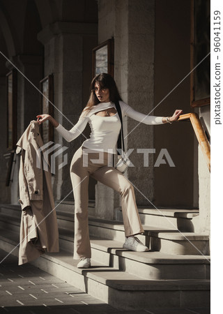 beautiful brunette girl dressed in white blouse, pants, sneakers with a handbag and camel coat in hands. Stylish trendy fashion outlook 96041159