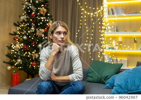 Sad and lonely young beautiful woman sitting at home on the sofa near the Christmas tree. Spends Christmas holidays alone. Sad and lonely young beautiful woman sitting at home on the sofa near the Christmas tree. Spends Christmas holidays alone. 96042907