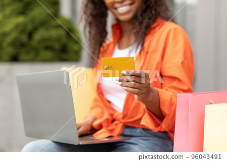 Smiling millennial black lady in casual with many bags typing on laptop, holding credit card in city, cropped 96043491