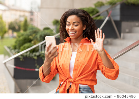 Happy young black lady has video call on phone, waving hand at video camera, enjoy conversation and gossip Happy young black lady has video call on phone, waving hand at video camera, enjoy conversation and gossip 96043496