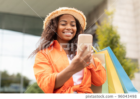 Smiling black lady with many packages with purchases has video call on phone, enjoys talk in city Smiling black lady with many packages with purchases has video call on phone, enjoys talk in city 96043498