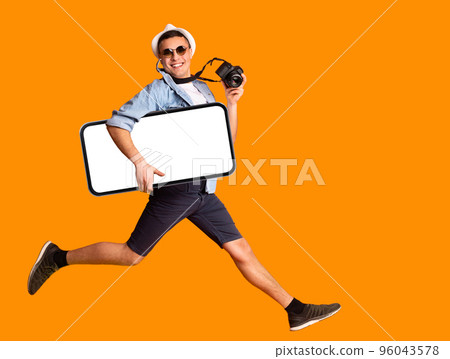 Cheerful Male Tourist Holding Camera Jumping With Big Blank Smartphone In Hand Cheerful Male Tourist Holding Camera Jumping With Big Blank Smartphone In Hand 96043578