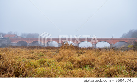 Kuldiga brick bridge over Venta in Kuldiga, Latvia on a foggy autumn day. Beautiful landscape with nature and old town architecture and bridge 96043743