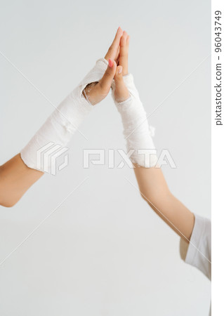 Vertical close-up cropped studio shot of unrecognizable injured woman and little kid girl with broken hand wrapped in plaster bandage giving high five at each, on white isolated background. 96043749