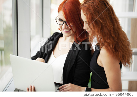 Two red-haired businesswomen in a co-working space with a laptop on the desktop in the office 96044003