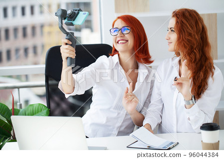 Two red-haired businesswomen in the office recording a video blog 96044384