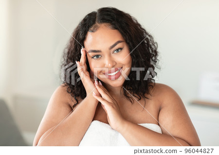 Portrait of beautiful african american chubby woman touching face while posing wrapped in towel and smiling at camera 96044827