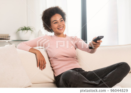 Smiling relaxed millennial african american woman with remote control watching tv sit on sofa, enjoy free time 96044829