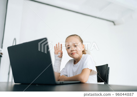 Positive little girl with broken hand wrapped in white plaster bandage showing hello gesture looking at camera sitting at desk with laptop in light room. Concept of child insurance and healthcare. Positive little girl with broken hand wrapped in white plaster bandage showing hello gesture looking at camera sitting at desk with laptop in light room. Concept of child insurance and healthcare. 96044887