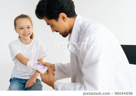 Friendly pediatrician male in uniform examining smiling little girl patient with broken hand wrapped in plaster bandage at checkup meeting, on white isolated background in professional studio. 96044896