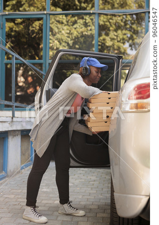 African american restaurant courier delivering pizza by car, side view. Pizzeria delivery service employee in headphones holding fastfood boxes stack, waiting for customer near company building African american restaurant courier delivering pizza by car, side view. Pizzeria delivery service employee in headphones holding fastfood boxes stack, waiting for customer near company building 96046547