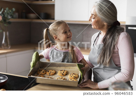 Smiling caucasian little girl and aged lady show fresh cookies on baking sheet, enjoy sweets in modern kitchen 96046583