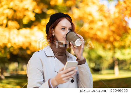 Cheerful caucasian millennial female with red hair in raincoat and hat, chatting by phone, drinking 96046665