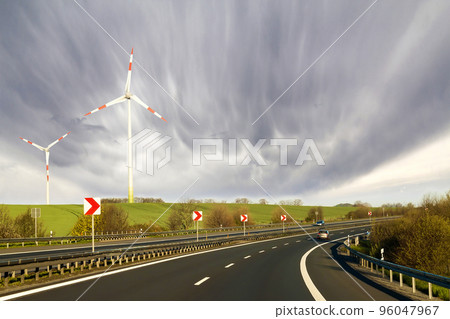 Wide highway with moving car stretching to horizon by green hills with high wind turbines on blue cloudy sky background. Modern technologies, renewable energy source and ecological problems solving. Wide highway with moving car stretching to horizon by green hills with high wind turbines on blue cloudy sky background. Modern technologies, renewable energy source and ecological problems solving. 96047967