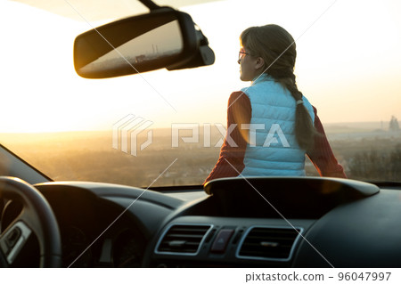 View from inside of a young woman standing near her car enjoying warm sunset view. Girl traveler leaning on vehicle hood looking at evening horizon. 96047997