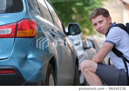 Sad young man driver sitting near his dented car looking shocked on damaged fender in road accident Sad young man driver sitting near his dented car looking shocked on damaged fender in road accident 96048136