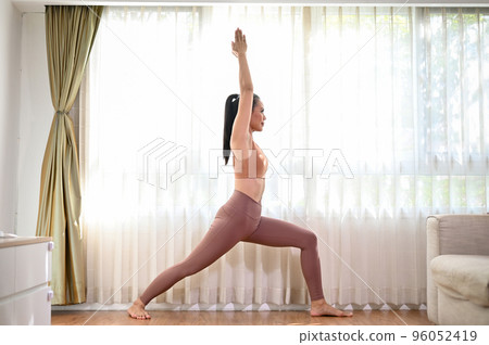 Beautiful Asian woman practicing yoga at home, practicing High Lunge pose or warrior I pose. 96052419