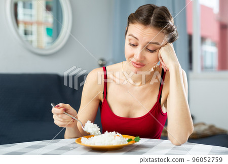 Sad woman eating rice while sitting at the table in the room. Diet concept and diet foods Sad woman eating rice while sitting at the table in the room. Diet concept and diet foods 96052759