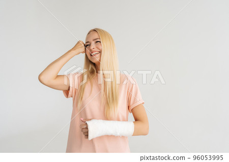 Studio portrait of cheerful young woman with broken arm wrapped in plaster bandage beating head with fist, standing on white isolated background, looking away. Concept of insurance and healthcare. Studio portrait of cheerful young woman with broken arm wrapped in plaster bandage beating head with fist, standing on white isolated background, looking away. Concept of insurance and healthcare. 96053995