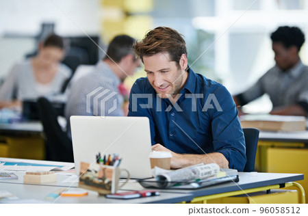 Hell tackle any project. Shot of a designer sitting at his desk working on a laptop with colleagues in the background. Hell tackle any project. Shot of a designer sitting at his desk working on a laptop with colleagues in the background. 96058512