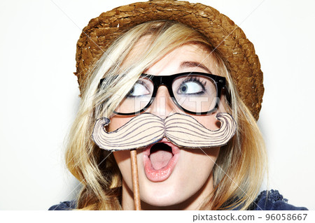 I moustache you a question.... Closeup of a young woman holding a moustache prop up to her face and looking surprised. 96058667