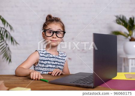 Cute little girl is sitting at table with her laptop and notebook, wearing glasses 96060175