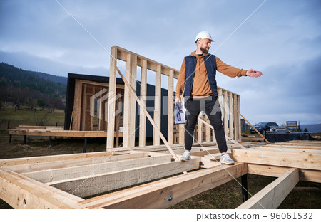 Male engineer building wooden frame house. Man builder standing on construction site in safety helmet, with construction documentation inspecting quality of work. 96061532