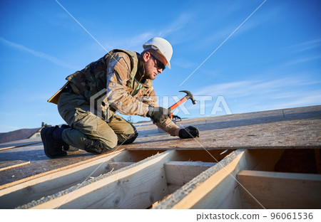 Carpenter hammering nail into OSB panel on the roof top of future cottage on blue sky background. Man worker building wooden frame house. Carpentry and construction concept. 96061536