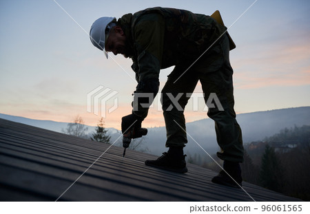 Male builder installing black corrugated iron sheet used as facade of future cottage. Silhouette of man worker building wooden frame house. Carpentry and construction concept. Male builder installing black corrugated iron sheet used as facade of future cottage. Silhouette of man worker building wooden frame house. Carpentry and construction concept. 96061565