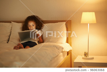 This is so relaxing. A cute little girl lying in a double bed and playing on a tablet. 96063068