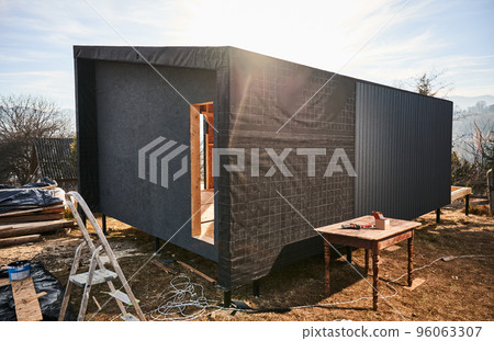 Wooden frame house on pile foundation in the Scandinavian style barnhouse under construction. Wall with waterproof membrane and black corrugated iron sheet used as facade of future cottage. 96063307
