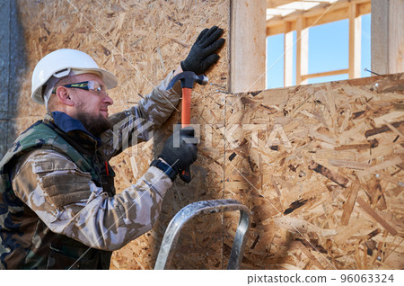 Carpenter hammering nail into OSB panel on the wall of future cottage. Man worker building wooden frame house. Carpentry and construction concept. 96063324