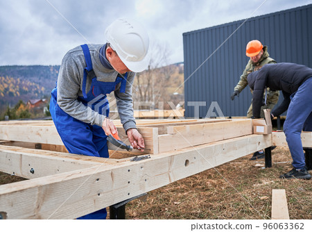 Man worker building wooden frame house on pile foundation. Carpenter hammering nail into wooden plank, using hammer. Carpentry concept. 96063362