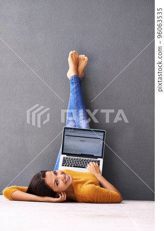 The up-side of technology. A young woman lying on the floor with her laptop against a gray background. 96063535