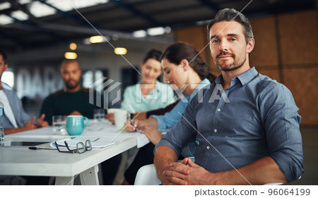 The creative process. Portrait of a mature man sitting at a table in an office with colleagues working in the background. The creative process. Portrait of a mature man sitting at a table in an office with colleagues working in the background. 96064199