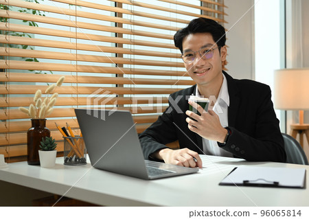 Successful businessman in formal suit sitting front of laptop at working desk and smiling to camera Successful businessman in formal suit sitting front of laptop at working desk and smiling to camera 96065814