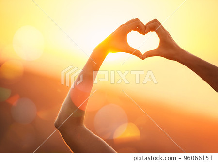 In love with the landscape. Shot of an unidentifiable womans hands making a heart shape over a sunset landscape. 96066151