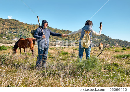 Back view of couple holding hands while walking on rural meadow with horses grazing on autumn or spring pasture. Woman takes a photo with smartphone. Unity with nature. Road to mountain. 96066430