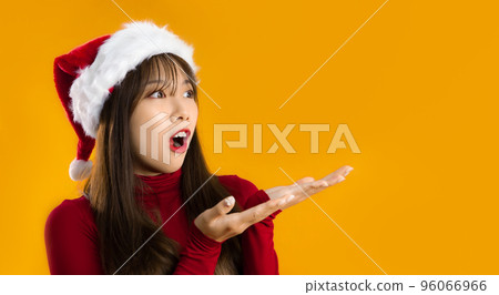 Beautiful asian woman in red sweatshirt wear santa hat hands pointing up yellow background. Merry Christmas theme. 96066966