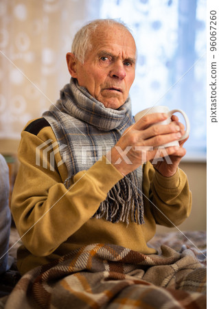 old man wrapped in blanket has cold and drinks hot infusion 96067260