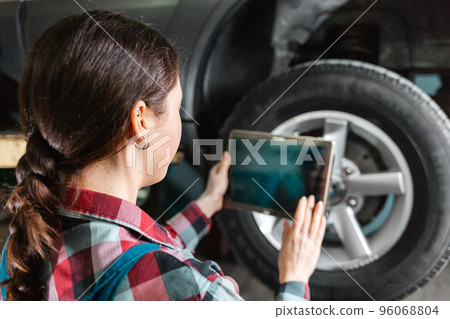 A young female mechanic in uniform with a tablet, conducting modern diagnostics. Car wheel in the background close up. Rear view A young female mechanic in uniform with a tablet, conducting modern diagnostics. Car wheel in the background close up. Rear view 96068804