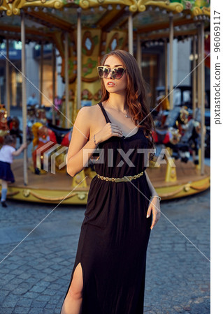 Portrait of a beautiful brunette woman in evening dress next to a children's carousel. 96069117