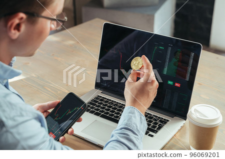 Cropped shot of crypto trader with phone and gold bitcoin at table with laptop, investing in btc Cropped shot of crypto trader with phone and gold bitcoin at table with laptop, investing in btc 96069201