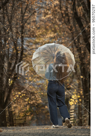 Brunette with long hair in blue sweater and pants stands with back under transparent umbrella in autumn park Brunette with long hair in blue sweater and pants stands with back under transparent umbrella in autumn park 96070297