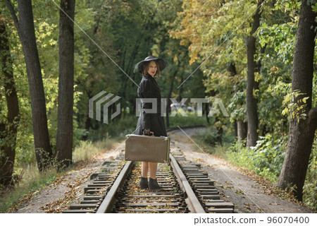 Woman in autumn fall forest walks along railway track with suitcase. Classic black coat and broad-brim hat. Back view Woman in autumn fall forest walks along railway track with suitcase. Classic black coat and broad-brim hat. Back view 96070400