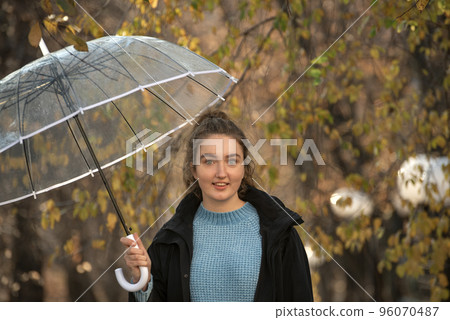 Charming girl with blue eyes looks into camera in autumn park with transparent umbrella. Walk in park 96070487