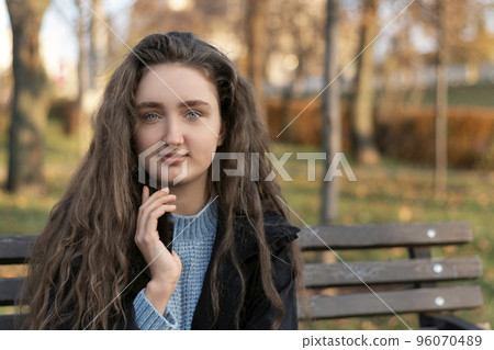 Beautiful young woman with long brown hair blue eyes and plump lips in autumn park on bench with phone in hands. 96070489