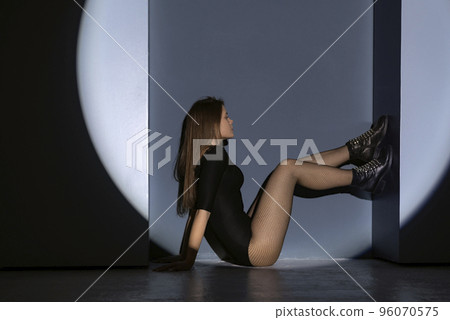 Sexy young model sitting and poses with feet resting on blue wall. Woman in projection of light from circular lamp. 96070575