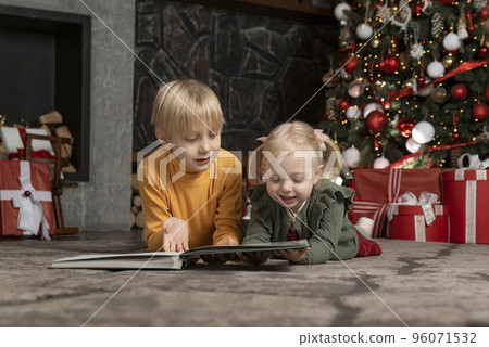 Two blond siblings lie on floor under Christmas tree. Smiling brother and sister reads the book. Christmas eve Two blond siblings lie on floor under Christmas tree. Smiling brother and sister reads the book. Christmas eve 96071532