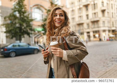 Young woman with curly blonde hair using the phone with a cup of coffee in hands on the city streets Young woman with curly blonde hair using the phone with a cup of coffee in hands on the city streets 96072761